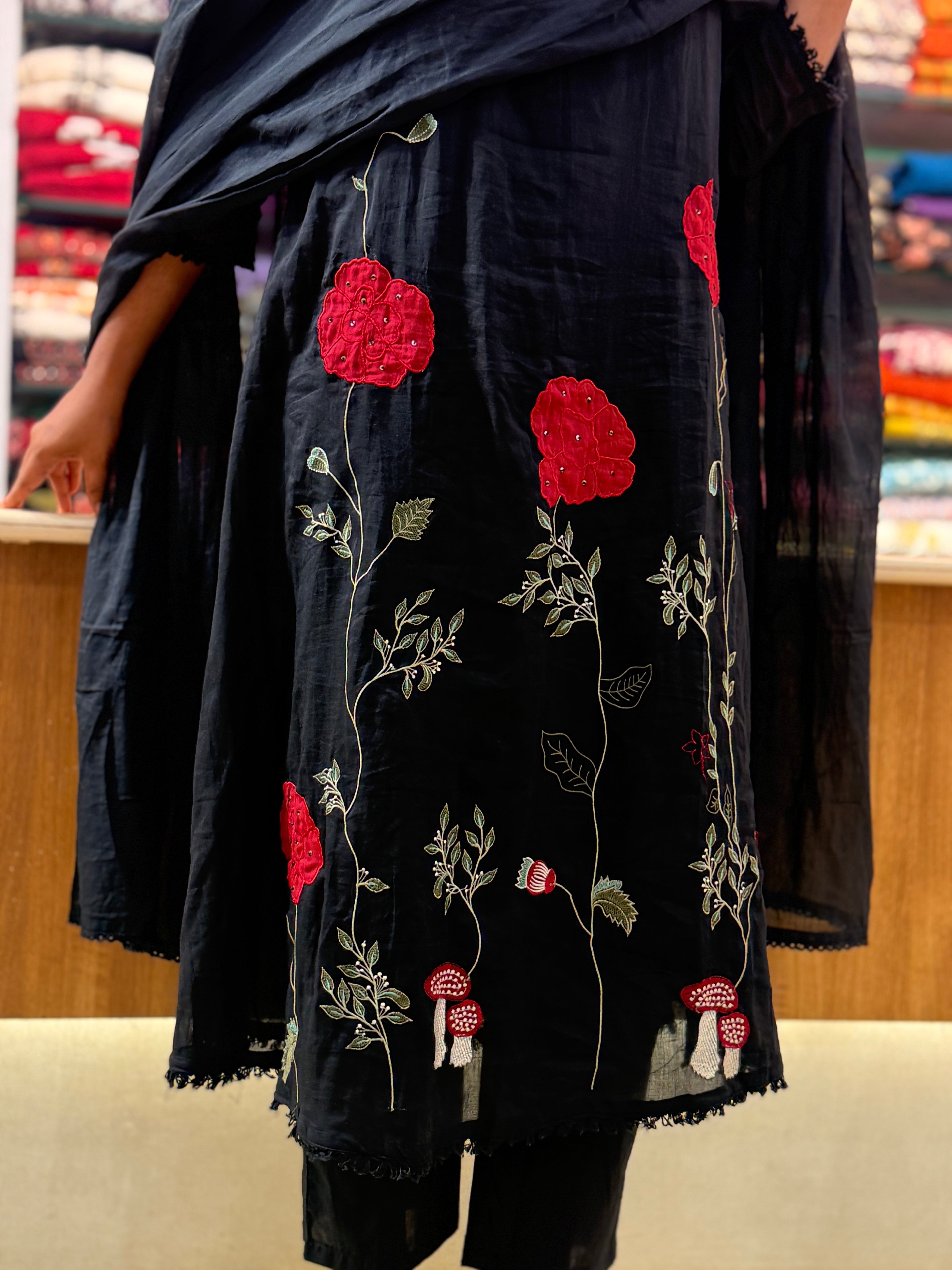 A black cotton kurta with red floral and green vine embroidery, paired with a matching black dupatta, shown on a model in a retail store.
